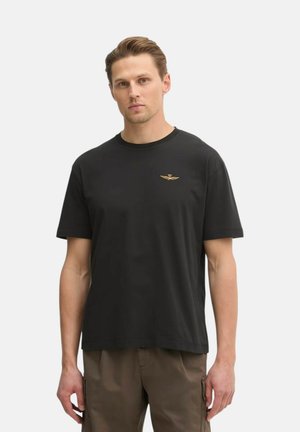 Uomo con capelli corti che indossa una maglietta nera con collo a giro e un piccolo emblema dorato alato sul petto, in piedi davanti a uno sfondo chiaro uniforme.