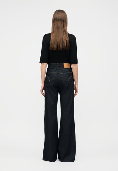 Γυναίκα με μακριά καστανά μαλλιά, φορώντας μαύρο κοντό τοπ και σκούρο, ψηλόμεσο, φαρδύ τζιν, στέκεται γυρισμένη με την πλάτη σε απλό φόντο.