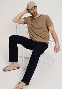 Jasnobrązowa koszulka, granatowe dżinsy, beżowe sandały. Siedzi na białym metalowym krześle, nosi beżową czapkę, w swobodnej i zrelaksowanej pozie.