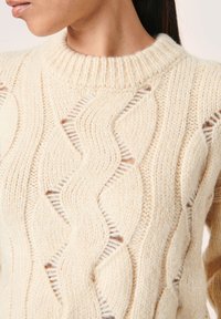 Suéter de punto color crema con cuello redondo acanalado, que presenta un patrón de ondas con detalles recortados y un acabado suave y texturizado.