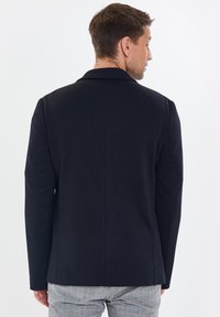 Blazer azul marino con un diseño estructurado, solapas con muescas y una textura suave. Ajuste tailored con líneas limpias y sin hardware ni acentos visibles.