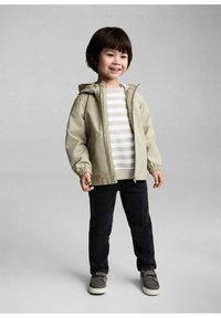 Giacca a vento verde chiaro con cappuccio, zip frontale, polsini elasticizzati. Camicia a righe beige e bianca sotto. Pantaloni scuri e scarpe grigie. Bambino che sorride.