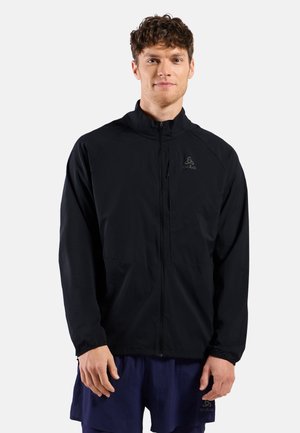 Jonge man met een zwarte jas met rits en marineblauwe korte broek, staand tegen een effen lichte achtergrond.