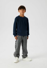 Pull en maille bleu marine à manches longues, associé à un jean en denim gris ample et des baskets blanches, se tenant sur un fond uni.