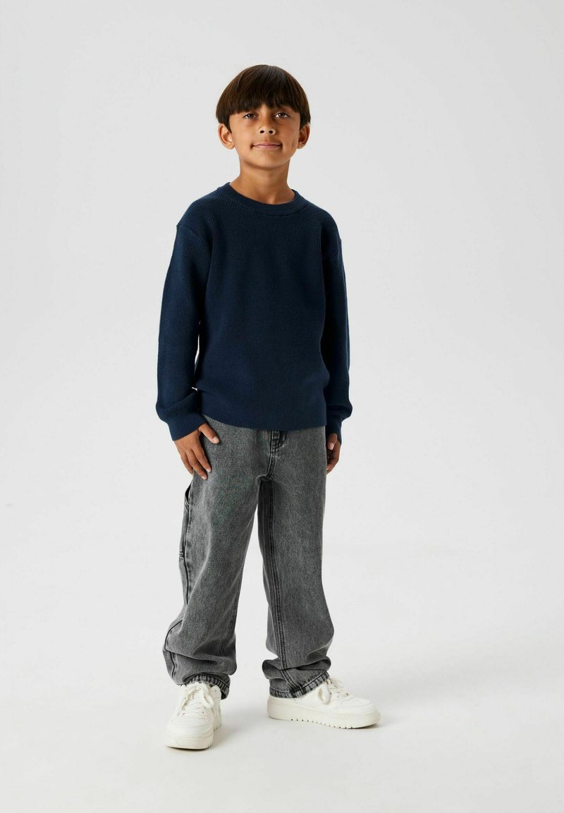 Pull en maille bleu marine à manches longues, associé à un jean en denim gris ample et des baskets blanches, se tenant sur un fond uni.