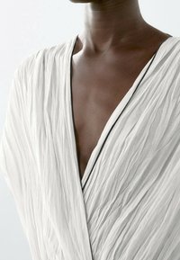 Personne à la peau foncée portant un vêtement blanc texturé avec un col en V profond bordé de noir, laissant apparaître le haut de la poitrine et le cou.