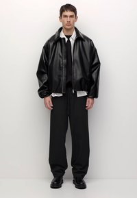 Jonge man die staat, wearing zwart faux leren jas, zwarte wijde broek, zwarte schoenen, witte shirt en zwarte sweater tegen een witte achtergrond.