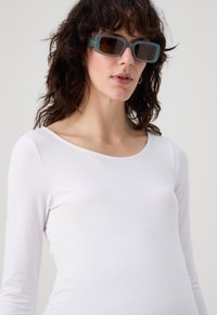 White long-sleeve top with rounded neckline. Model wears dark rectangular sunglasses with a teal blue frame and brown tinted lenses.