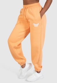 Orange sweatpants lavet af blødt stof, med elastisk talje, sidelommer og et hvidt sommerfuglemotiv på venstre lår.