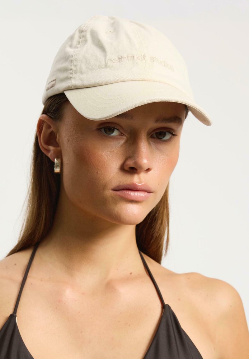 Cappellino da baseball beige con visiera curva e testo ricamato. Il modello indossa un top scuro, con semplici orecchini d'argento visibili. Tessuto morbido.