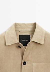 Veste en daim beige avec boutons noirs, un col et une étiquette noire visible portant la mention Massimo Dutti.