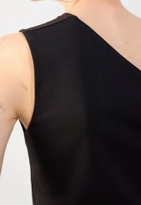 Gros plan d'une personne portant un haut noir sans manches avec une large bretelle, montrant une partie du haut du dos et du bras.