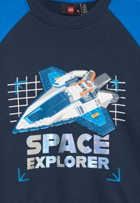Graphic di un'astronave LEGO blu e bianca sopra il testo riflettente "SPACE EXPLORER" su una maglietta raglan blu navy e blu.