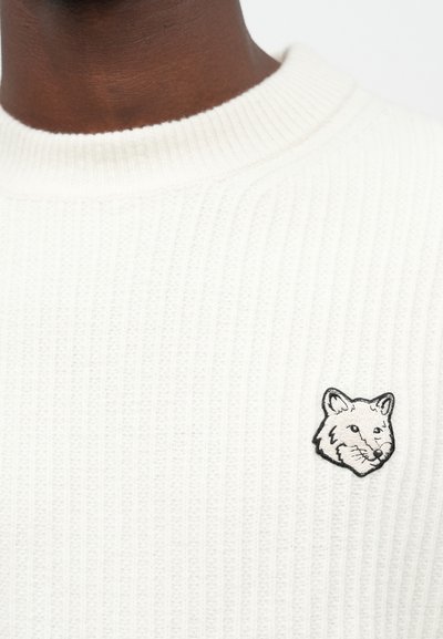 Close-up of a white ribbed sweater with a black and white embroidered fox head on the chest.