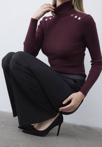 Bordeaux geribbelde coltrui met gouden knoopaccenten op de schouders, gecombineerd met zwarte wijde broek en zwarte stilettohakken.