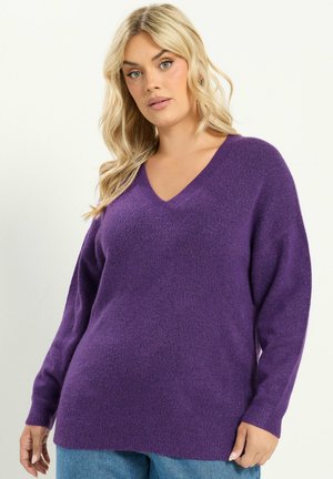 Femme blonde portant un pull violet à manches longues avec col en V et un jean bleu, debout devant un fond clair uni.