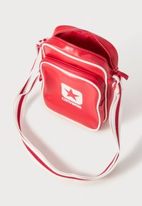Borsa Converse rossa e rettangolare realizzata in materiale liscio, con dettagli bianchi e cerniere. Include una tracolla a strisce bianca e rossa.
