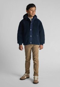 Marineblauwe fleecjas met een capuchon, drukknoopsluiting en geribbelde manchetten, gecombineerd met lichtbruine broeken en beige sneakers met witte accenten.