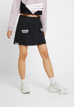 Personne portant une jupe noire Adidas avec zip devant et écussons, un top court, des chaussettes blanches et des baskets blanches, debout devant un fond uni.