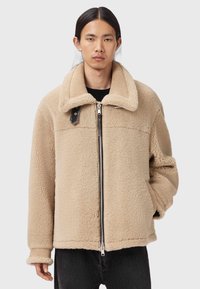 Beżowa polarowa kurtka z dużym kołnierzem, przednim zamkiem i czarnymi akcentami. Posiada teksturowany materiał i luźny krój.