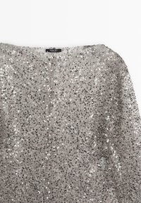 Top con paillettes argentate, dalla vestibilità ampia, con scollatura rotonda e tessuto trasparente. Numerosi piccoli e lucenti paillettes riflettono la luce.