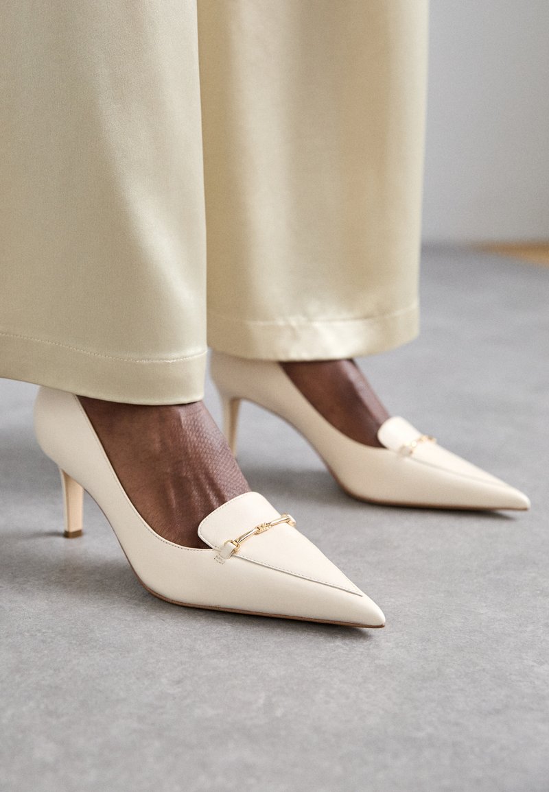 Escarpins beige à bout pointu en cuir lisse, ornés d'une chaîne dorée et d'un talon aiguille fin. Associés avec un pantalon crème.