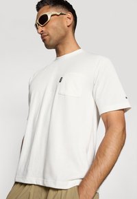 T-shirt bianco in cotone con maniche corte, taschino sul petto e una piccola etichetta nera sulla tasca. Texture liscia, vestibilità comoda.