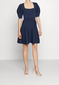 Námořnické modré šaty s balónovými rukávy a čtvercovým výstřihem, přiléhavý pas, rozšířená sukně, obuty se světlými sandály na blokovém podpatku se řemínky.