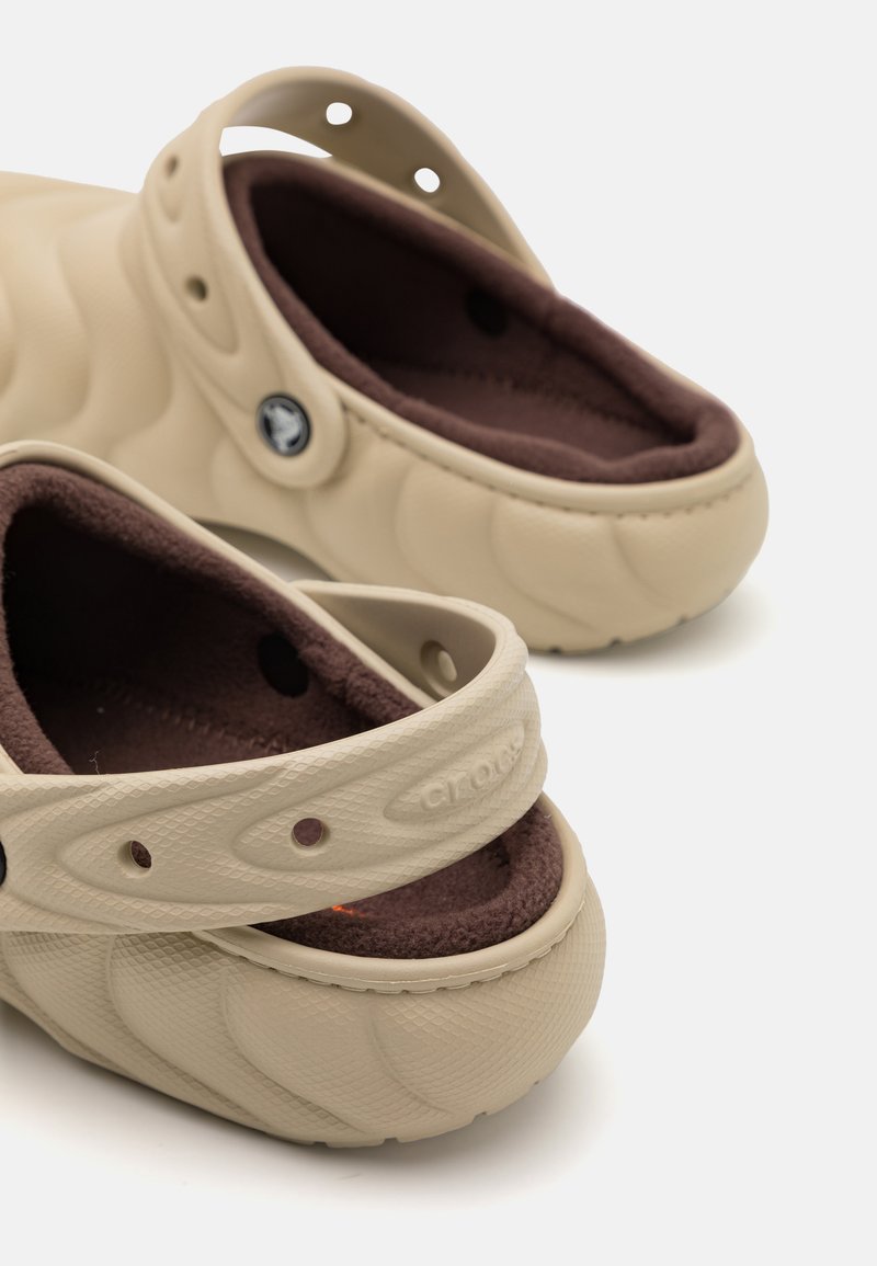 Des Crocs beiges en matériau semblable au caoutchouc, avec un extérieur texturé, une doublure en fausse fourrure marron, une sangle arrière et des trous de ventilation.