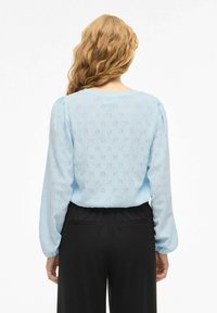 Lyseblå bluse med lange ermer, dekorert med broderte prikkmønstre og rund halsringning. Kombinert med svarte bukser, glatt stofftekstur.