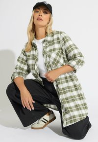 Zielona i biała koszula w kratę flanelową z podwiniętymi rękawami, noszona na białym t-shircie. Czarne spodnie o szerokich nogawkach i beżowe adidasy dopełniają stylizację.