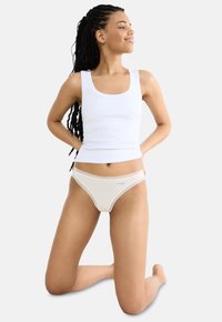 Top en coton blanc et thong couleur crème avec des détails en dentelle. Le mannequin est posé à genoux, mettant en valeur le tissu et la coupe.