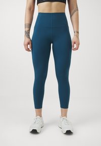 leggings teal a vita alta realizzati in materiale elasticizzato, caratterizzati da una texture liscia e una forma aderente, abbinati a scarpe da ginnastica bianche.
