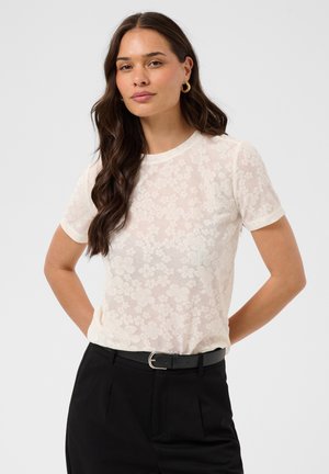 Femme aux longs cheveux bruns portant un haut blanc transparent à fleurs à manches courtes, un pantalon noir ceinturé et des boucles d’oreilles dorées, debout les mains derrière le dos.