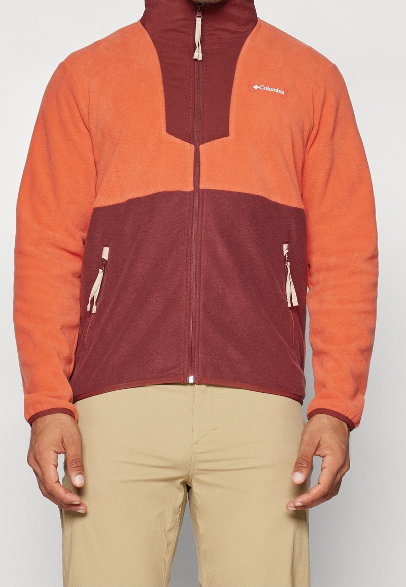 Giacca in pile con un design color block in arancione e bordeaux, colletto alto, chiusura a cerniera e tasche laterali. Texture liscia.