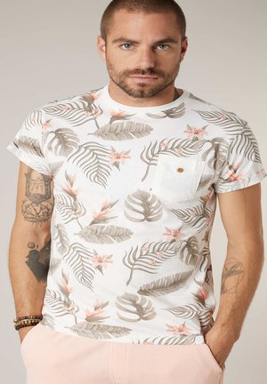 Homme aux cheveux courts et à la barbe, portant un t-shirt blanc à motifs floraux avec une poche et un pantalon rose clair, affichant des tatouages sur les deux bras.