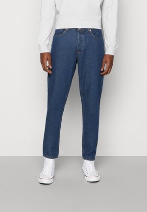 Jeans de ganga azul com corte reto, design de cinco bolsos e detalhes de costura subtil. Usados com sapatilhas brancas.
