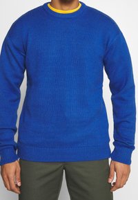 Niebieski sweter wełniany z okrągłym dekoltem, ściągaczami na mankietach i u dołu; noszony na żółtej koszuli, wykonany z teksturowanej, średniej grubości tkaniny.