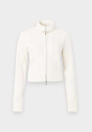 Pull en maille torsadée blanche, raccourci, avec un col montant et une fermeture éclair à l'avant. La texture est épaisse, avec des poignets et un ourlet côtelés.