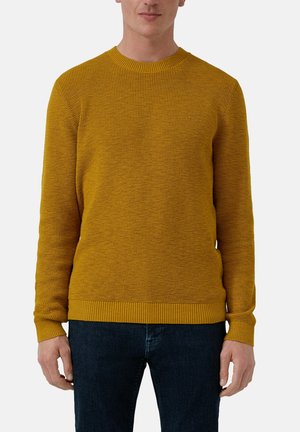 Mand iført en sennepsgul tekstureret striktrøje og mørkeblå jeans, stående mod en ensfarvet lys baggrund.