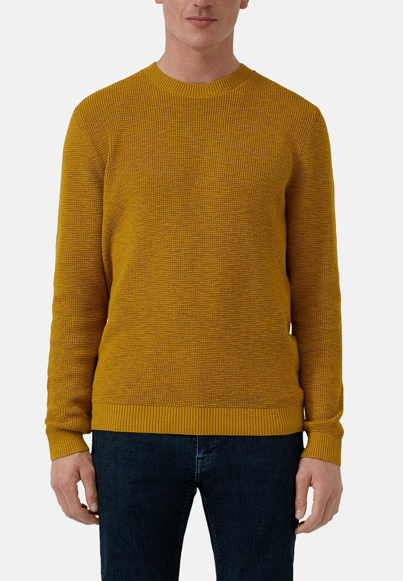 Homme portant un pull tricoté texturé jaune moutarde et un jean bleu foncé, debout devant un fond clair uni.