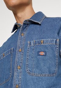 Camicia in denim di colore blu medio, con due tasche frontali, bottoni dorati e un patch con logo Dickies sulla tasca sinistra.