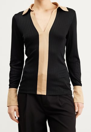 Femme portant un haut noir côtelé à manches longues avec col beige, patte de boutonnage devant et poignets, associé à un pantalon noir.