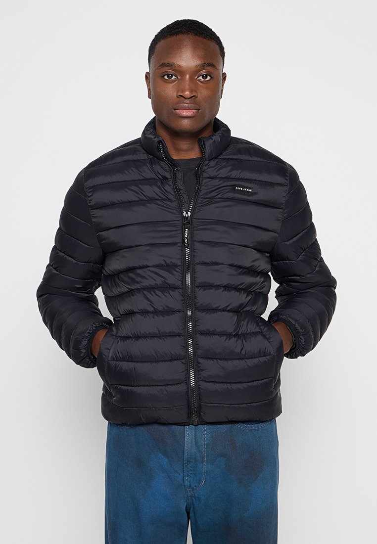 Veste doudoune noire avec fermeture éclair, design matelassé et col haut. Portée par-dessus une chemise sombre, associée à un jean bleu. Texture lisse.