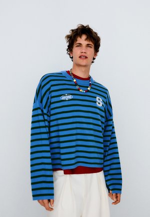 Junge Person mit lockigem Haar, die ein blau-schwarz gestreiftes Langarmshirt, weiße Hose und eine bunte Perlenkette vor einem schlichten Hintergrund trägt.