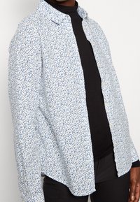 Camicia button-up bianca con motivo floreale blu, a maniche lunghe e colletto. Realizzata in tessuto leggero, presenta un orlo leggermente curvo.