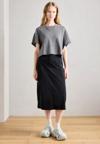 Woman wearing gray cropped top, black midi skirt with pockets, white socks, and chunky light gray slip-on shoes, standing on wood floor.