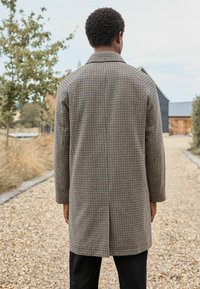 Ruudullinen takki kauluksella, valmistettu kankaasta, jossa on beige ja musta houndstooth-kuvio. Pitkät hihat ja takana halkio.