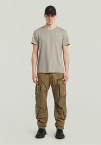 Jasnoszara bawełniana koszulka, beżowe spodnie cargo z wieloma kieszeniami i zamkami, czapka w kolorze czarnym oraz czarne buty. Casualowy styl uliczny.