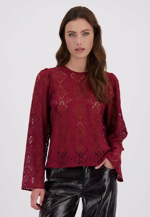 Vrouw met lang bruin haar, gekleed in een bordeauxkleurige kantblouse met opgeblazen mouwen en zwarte glanzende leren broek, staande tegen een witte achtergrond.